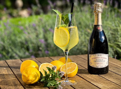 Auf einem Tisch im Grünen steht ein Limoncello Spritz, daneben Zitronen und eine Flasche Doppio Passo Prosecco